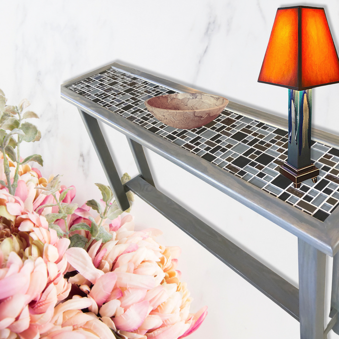 A grey tiled top console table with a handmade ceramic lamp with orange shade as well as a wooden salad bowl displayed on top.